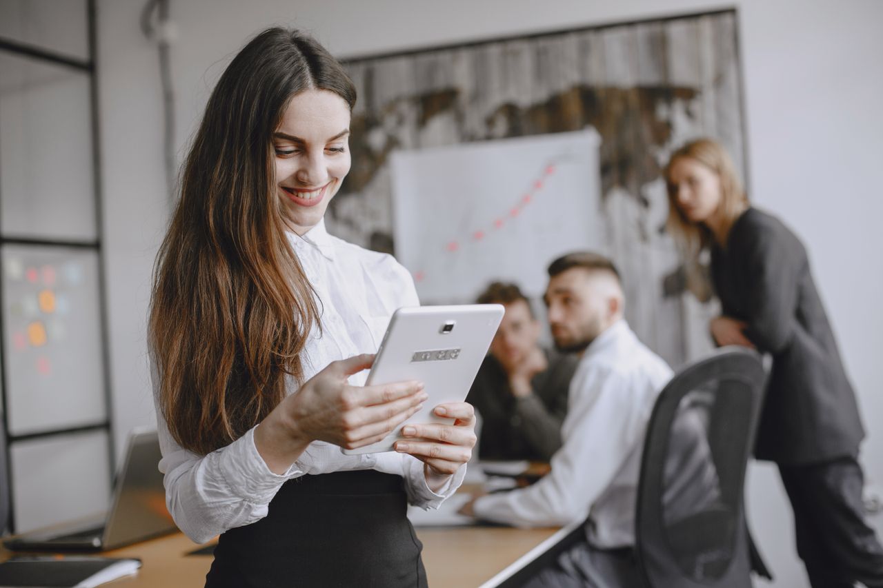 Mastering the First Impression: Your intriguing post title goes here Young woman in a professional office setting browsing a tablet with colleagues in the background.